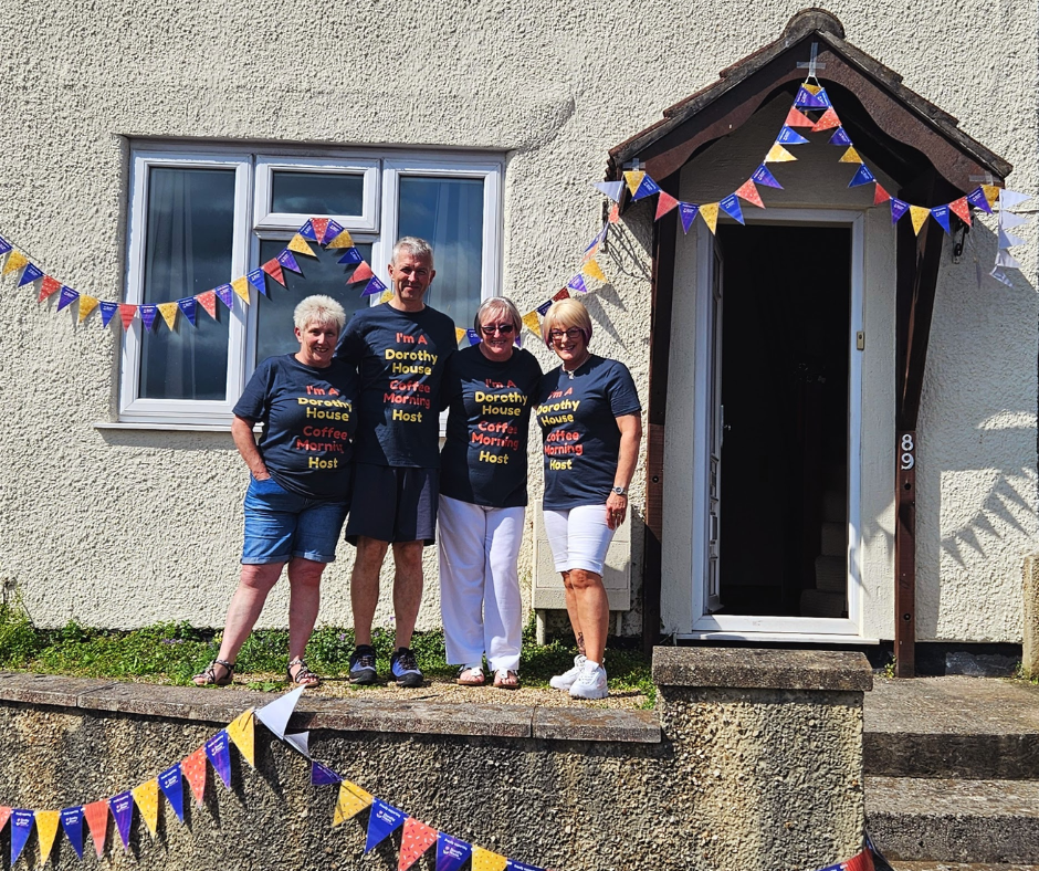 Three women and a man stand in front of a house which is decorated with blue, yellow and orange bunting. They are all smiling and wearing blue t-shirts that say "I'm a Dorothy House Coffee Morning host"