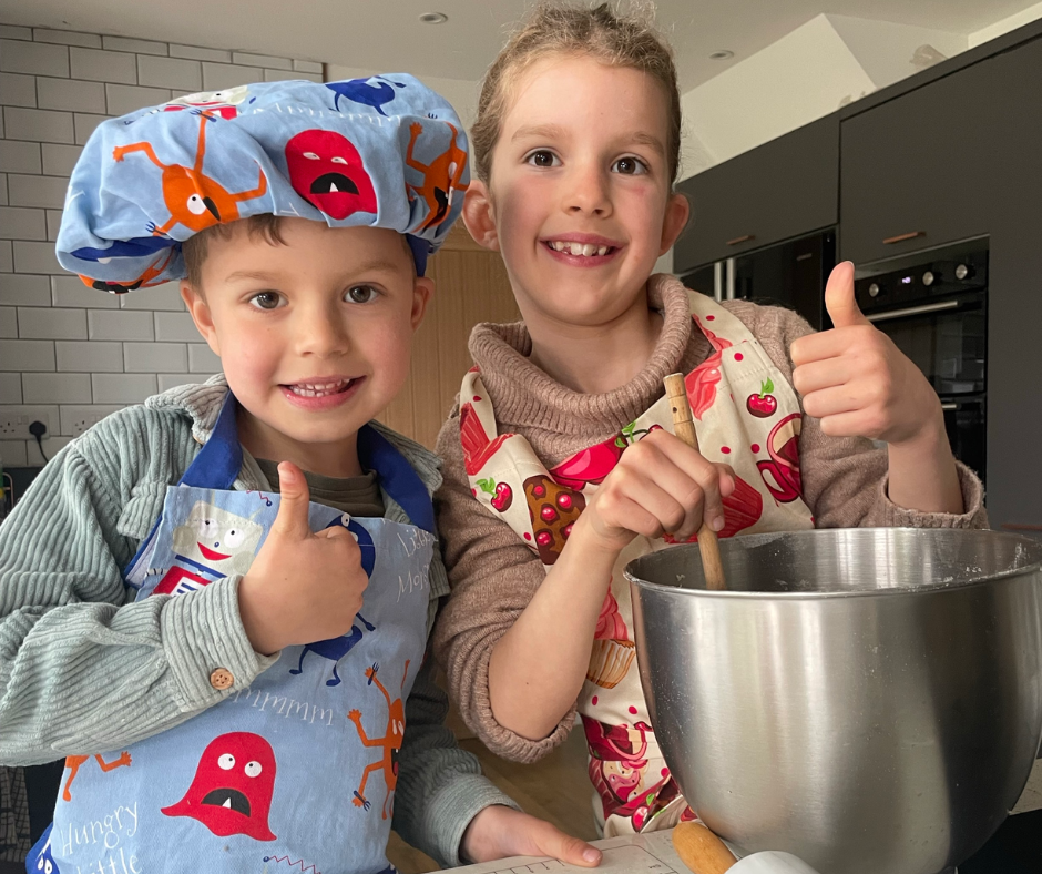 A young boy dressed in a grey top, blue apron and matching chef's hat, covered with colourful monsters. He is stood next to a young girl who is wearing a pink jumper and a pale yellow apron patterned with red cupcakes. The two children are stood next to a mixing bowl, both smiling and giving a thumbs up.