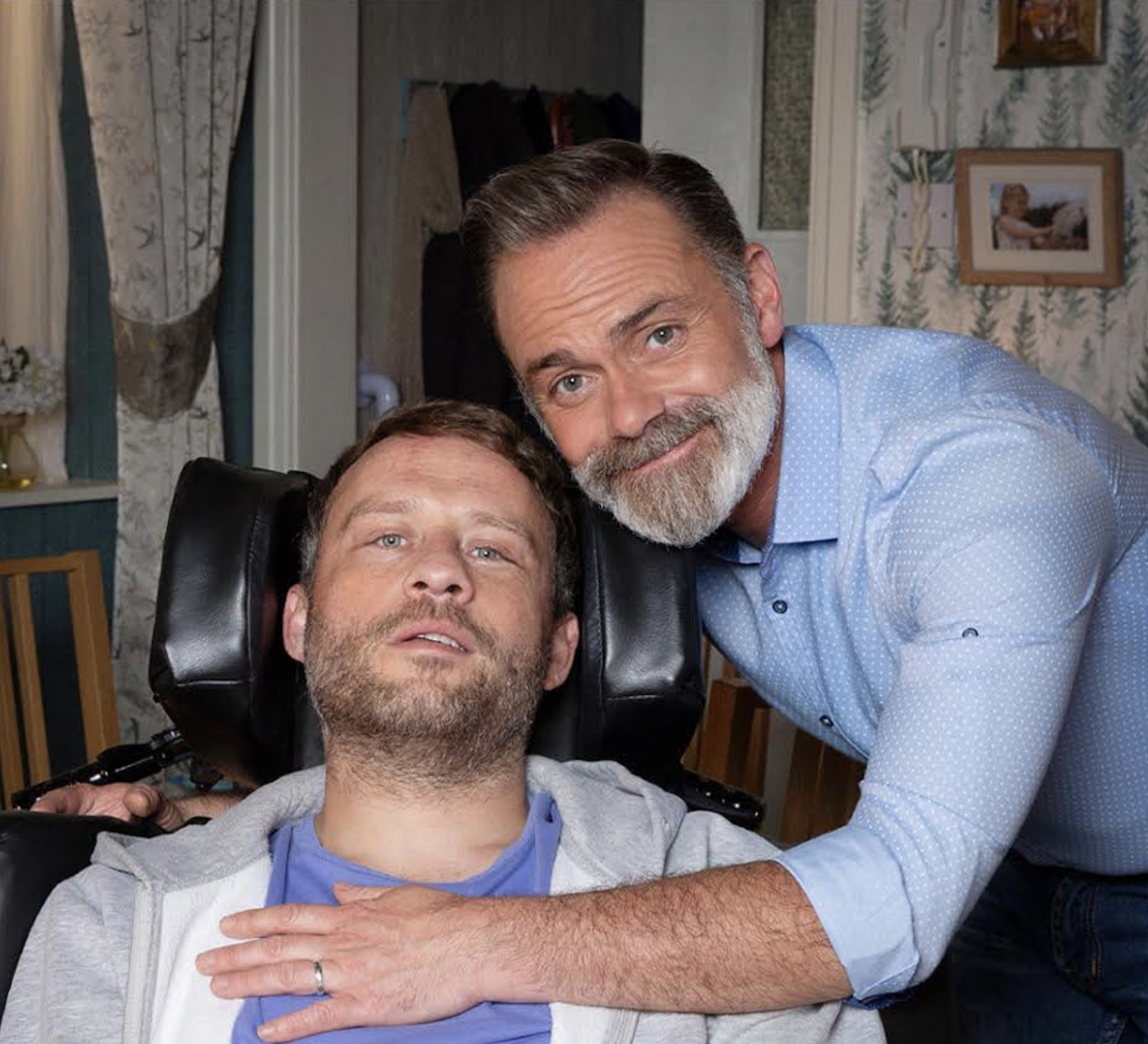 A man with a beard, wearing a blue shirt puts his arm around a man in a wheelchair. The man is wearing a blue shirt and grey hoodie.